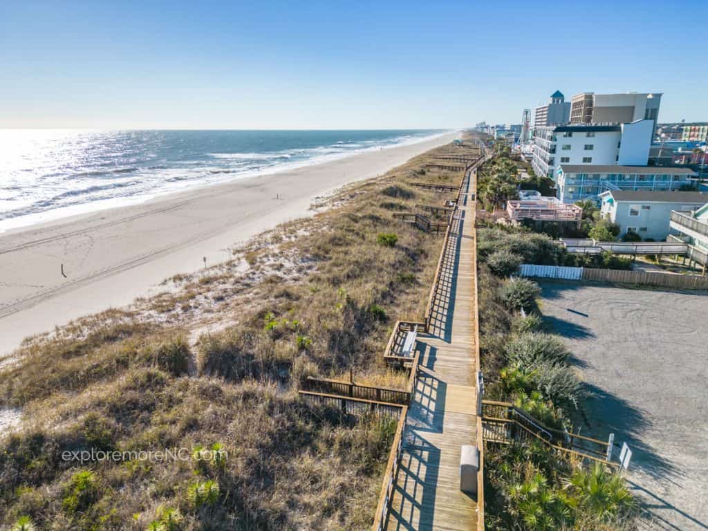A perspective of Carolina Beach from a wooden boardwalk, showing the natural dune landscape leading to a line of beachfront buildings under a clear sky.