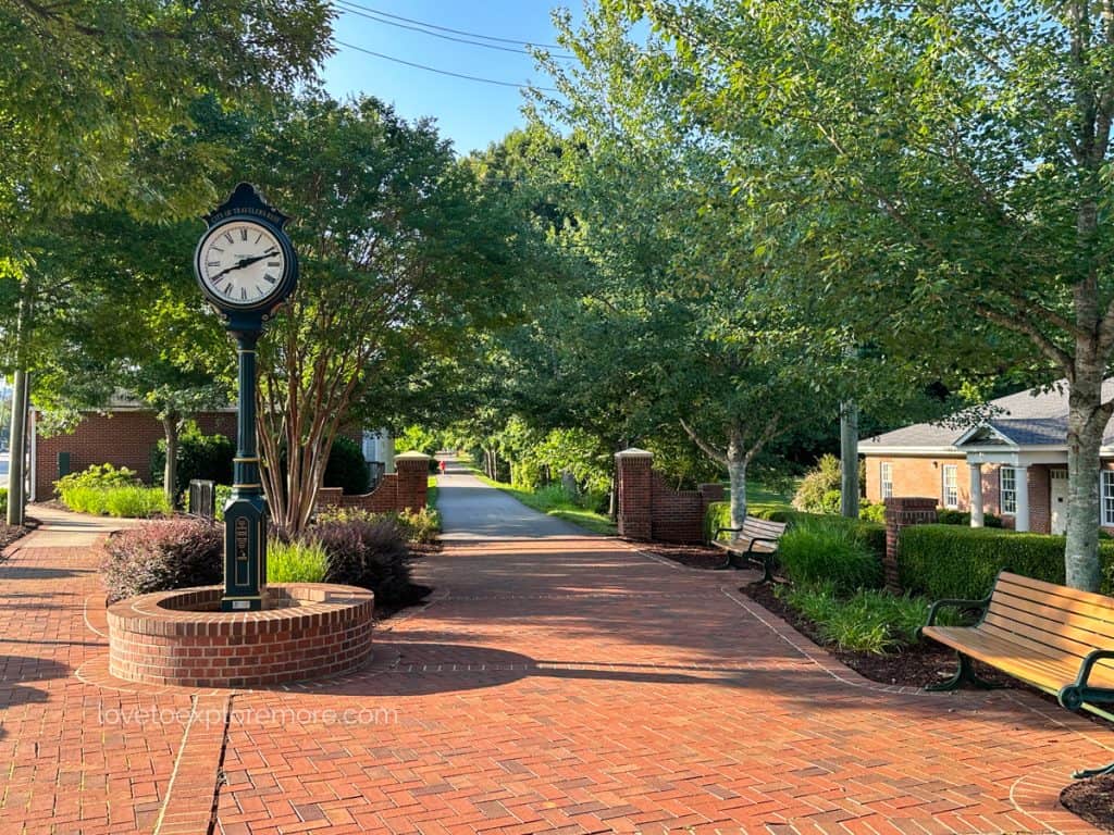 Charming downtown Travelers Rest with a classic street clock, brick pathways, benches, and lush greenery.