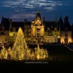 A stunning view of the Biltmore Estate at night with a large Christmas tree illuminated by twinkling lights in the foreground.