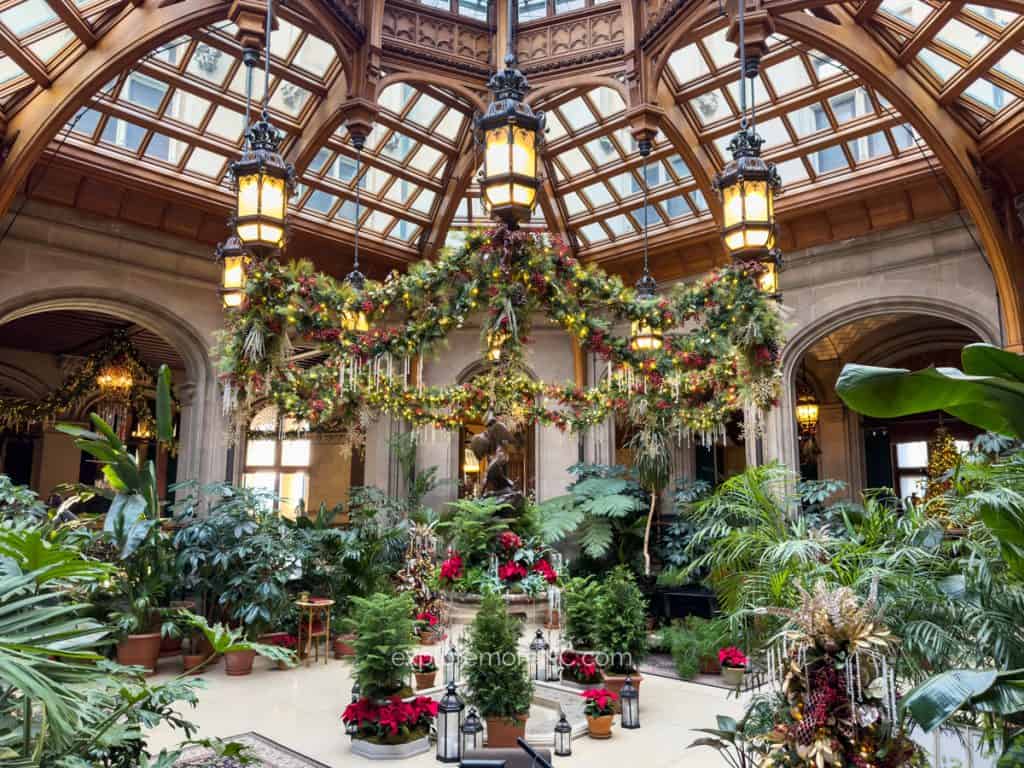 The Biltmore Estate conservatory decorated for Christmas with hanging garlands, festive greenery, and warm lighting under its glass ceiling.